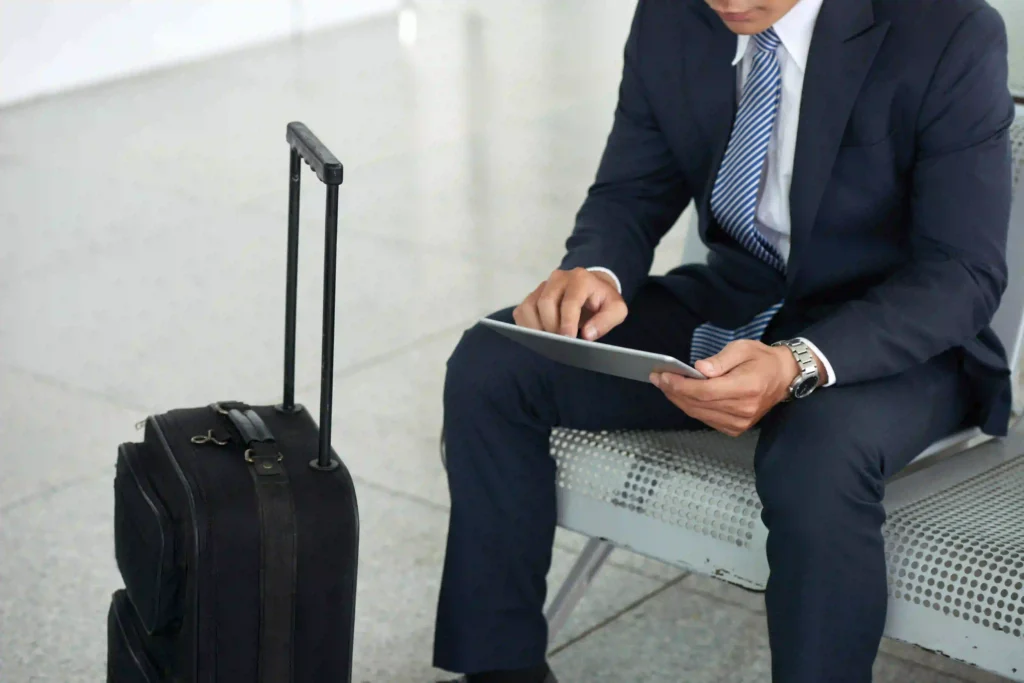 Person sitting on a bench at the airport, wearing a formal suit and using a tablet. Next to them, a black trolley with extended handle resting on the shiny floor.