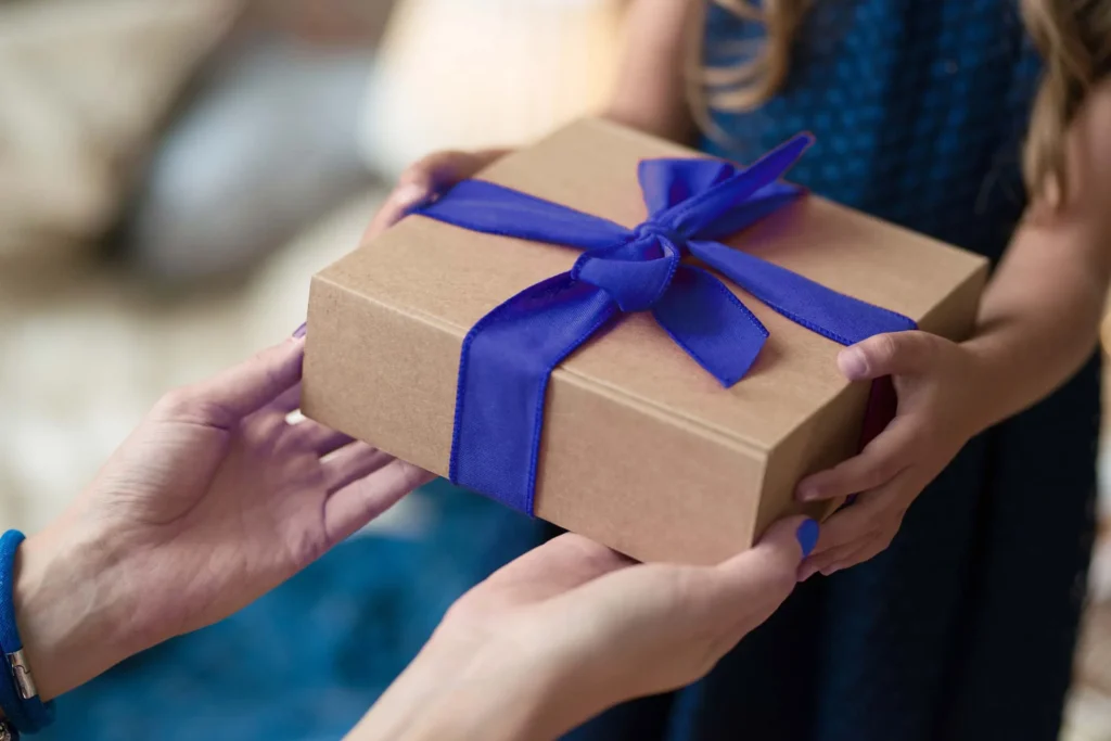 Hands giving and receiving a rectangular gift box wrapped in brown paper, decorated with a bright blue ribbon. In the background, a blue outfit and a blurred indoor setting are visible
