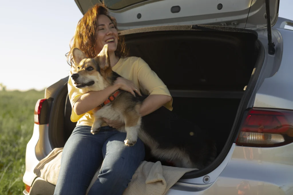 Person sitting on the edge of an open car trunk, hugging a Corgi dog. The dog faces outward, with green grass and natural light in the background
