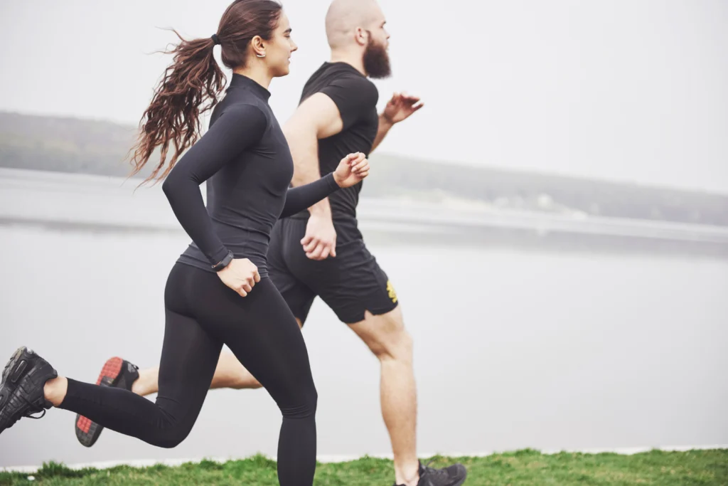 Two people running outdoors along a grassy trail near a river. They are wearing dark sportswear and running shoes, with natural scenery and water in the background