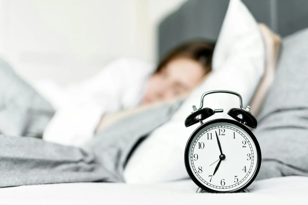Close-up of a black analog alarm clock with white dial, placed on a bed with gray sheets and white pillows. In the background, a blurred figure lying down in a bright room.