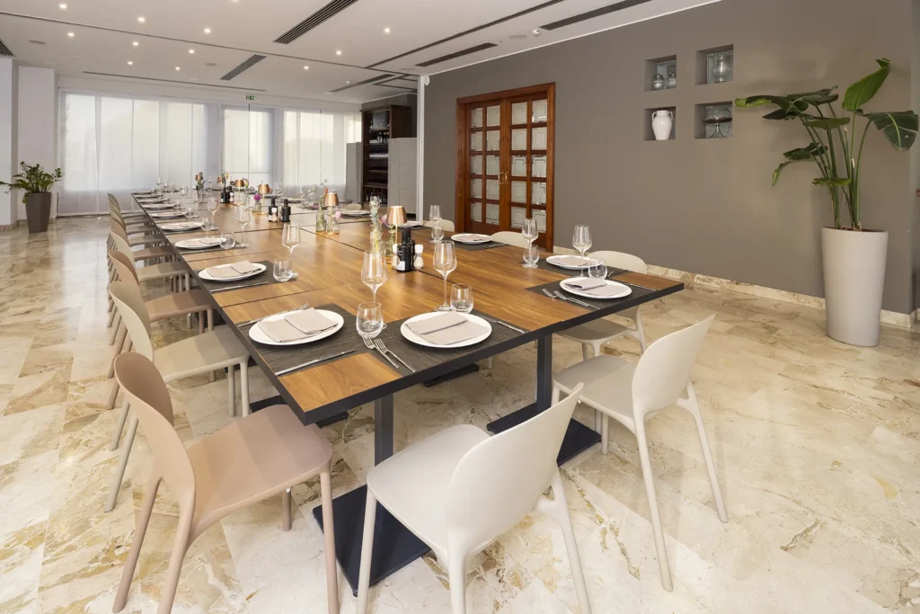 Elegant room with light marble flooring and grey walls. In the center, a long rectangular wooden table set with white plates, wine and water glasses, cutlery, and dark placemats. Modern light-colored plastic chairs line the sides. On the table, bottles of wine and minimal decorations. In the background, a wooden door with glass panels and a large potted plant.